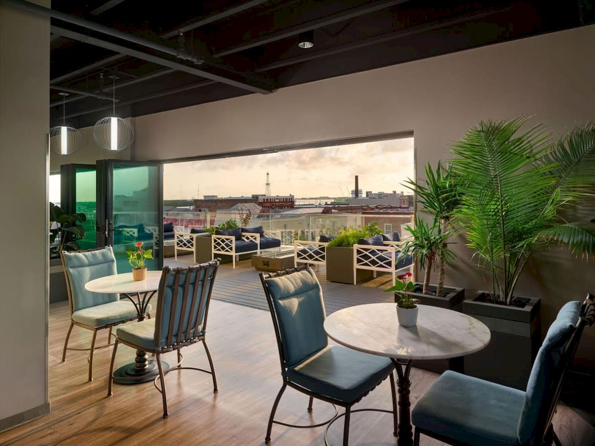 The image shows a stylish indoor seating area with round tables and chairs, opening to a balcony with outdoor seating and a city view.
