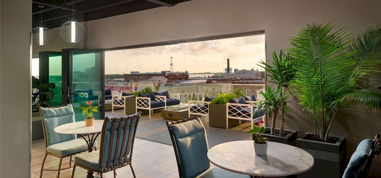 The image shows a stylish indoor seating area with round tables and chairs, opening to a balcony with outdoor seating and a city view.