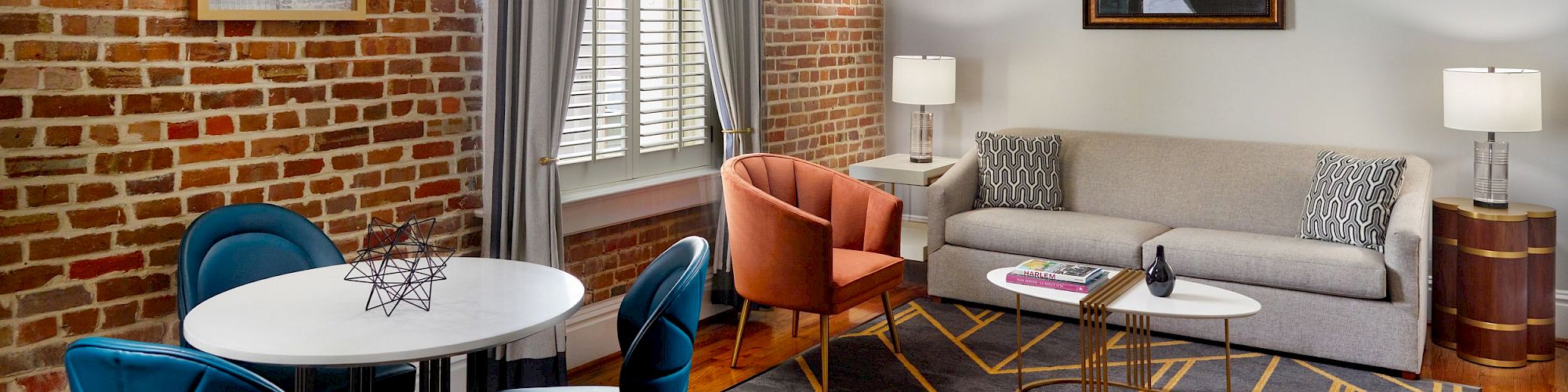 A modern living room with a brick wall, grey couch, orange chair, round tables, blue dining chairs, lamps, artwork, and a window with grey curtains.