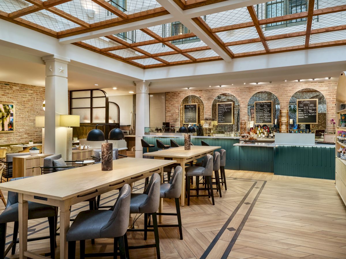 This image shows a modern cafe with wooden tables, cushioned chairs, a counter with a barista area, and a decorative ceiling with skylights.