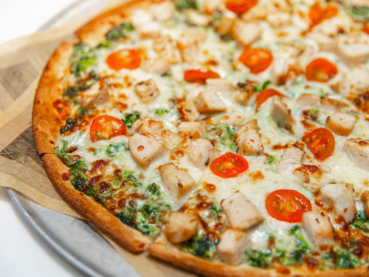 Wood-fired pizza with mozzarella, cherry tomatoes, chicken, and spinach on a parchment-lined tray.