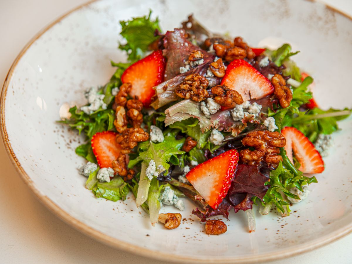 A bowl of mixed green salad with sliced strawberries, walnuts, and crumbled cheese on top, served in a white plate.