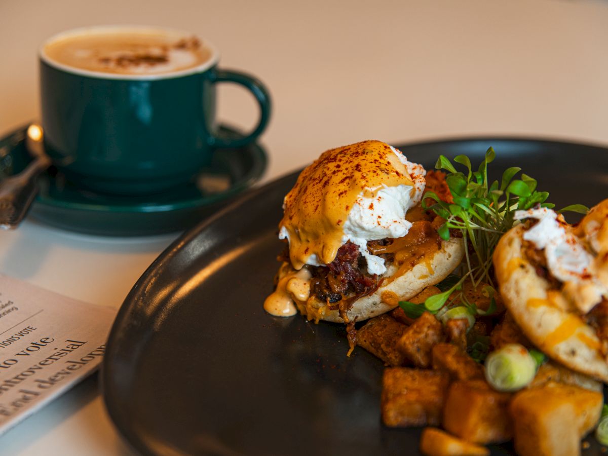 Eggs Benedict on a dark plate with greens and a drizzled sauce, alongside a cup of coffee in the background.