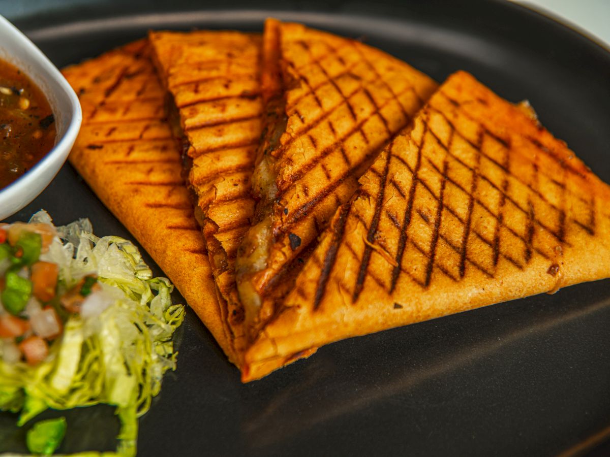 A grilled flatbread sandwich with crispy grill marks, served with a small cup of sauce and a side salad, on a plate.