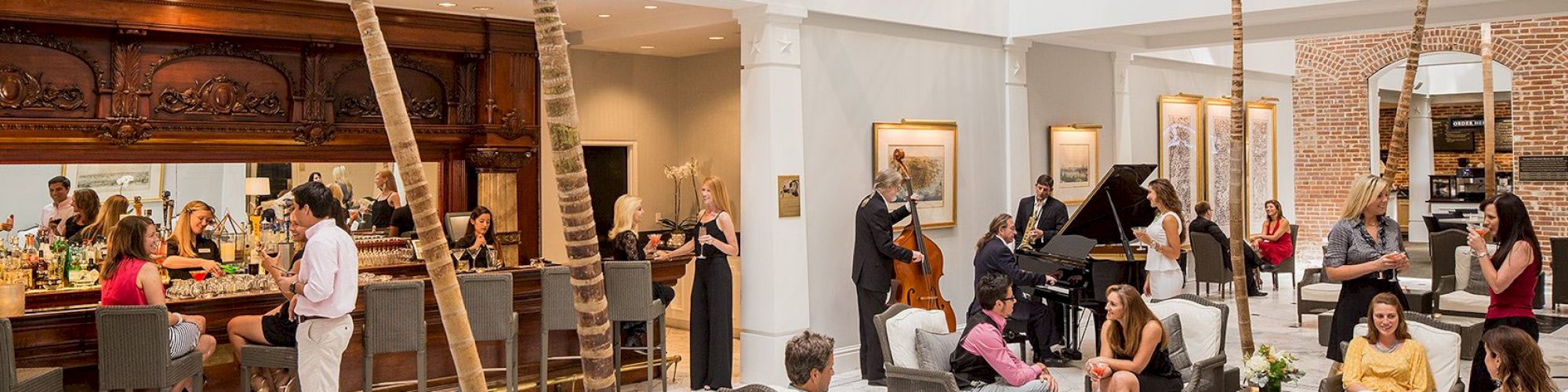 People are socializing in an elegant lounge with a bar, palm trees, and a live jazz band. Some are seated, while others chat at the bar.