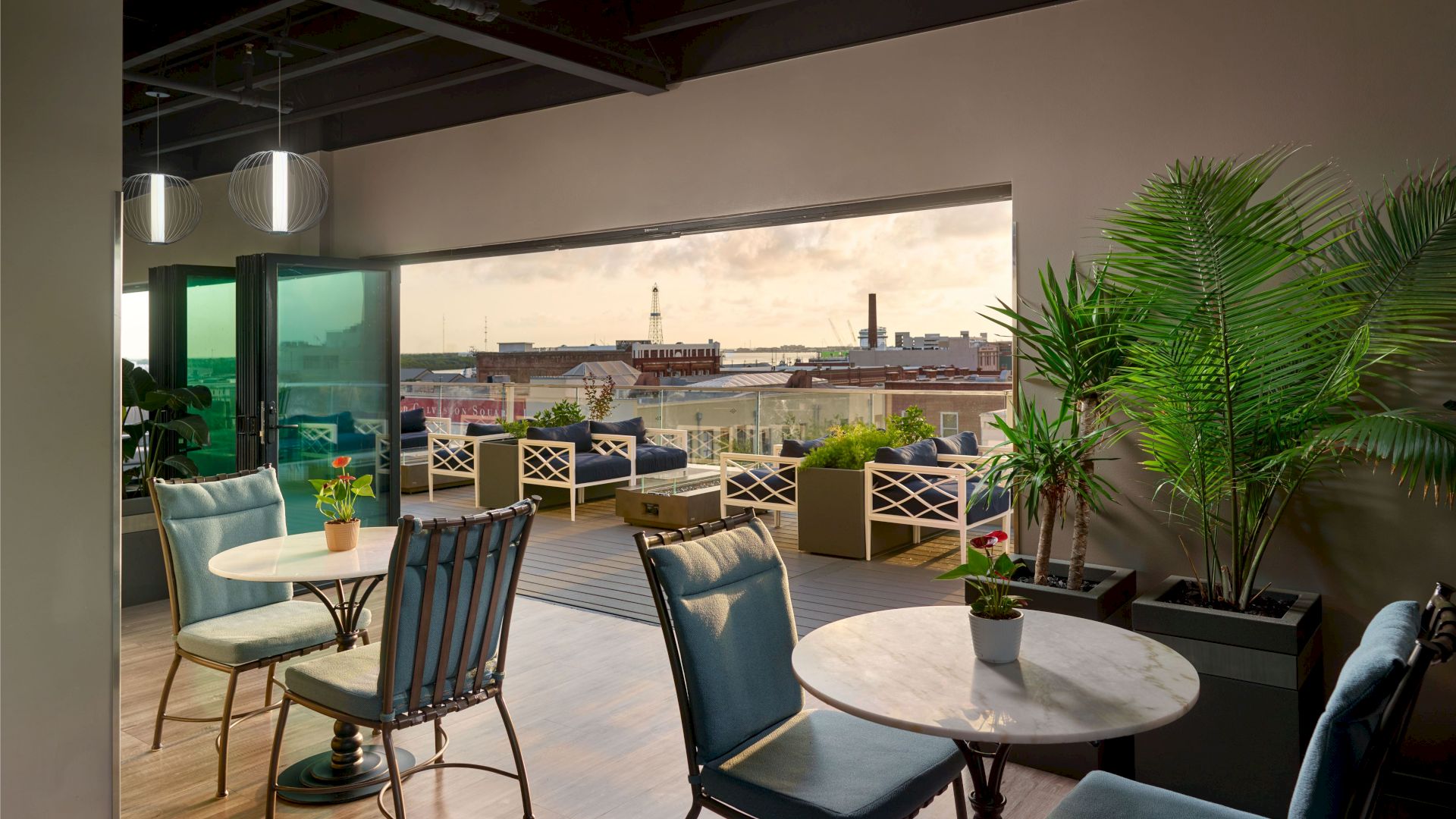 This image shows a modern indoor lounge area with tables, chairs, plants, and a view of an outdoor terrace overlooking a cityscape.