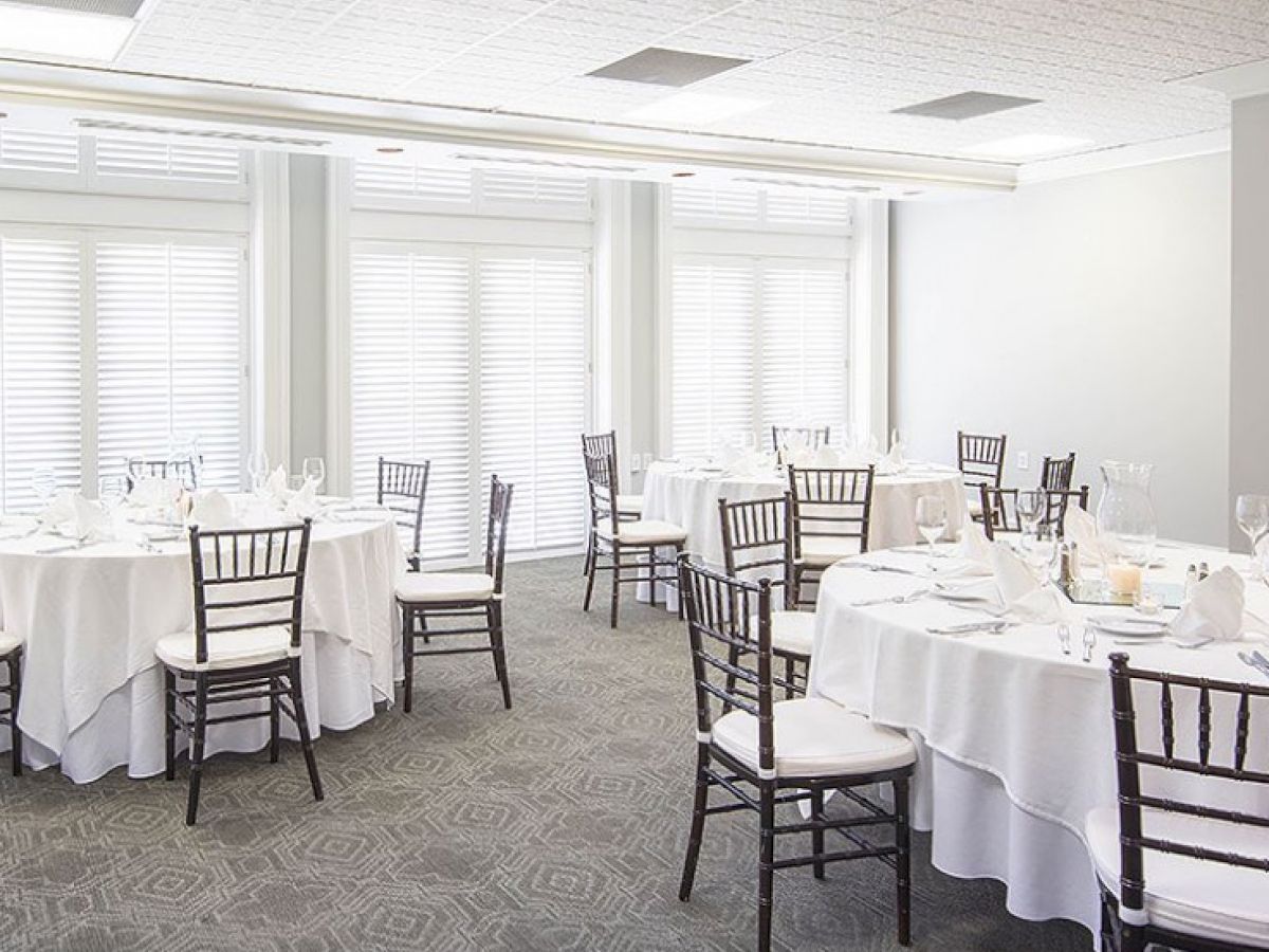 A well-lit banquet hall with several round tables covered with white tablecloths, set for a formal event with cutlery and glasses.