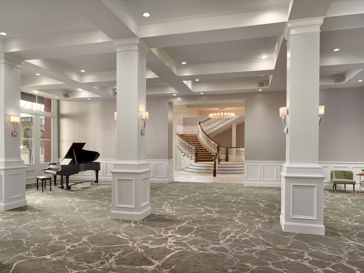 A spacious, elegantly decorated lobby or reception area with a grand piano, patterned carpet, modern lighting, and a staircase in the background.