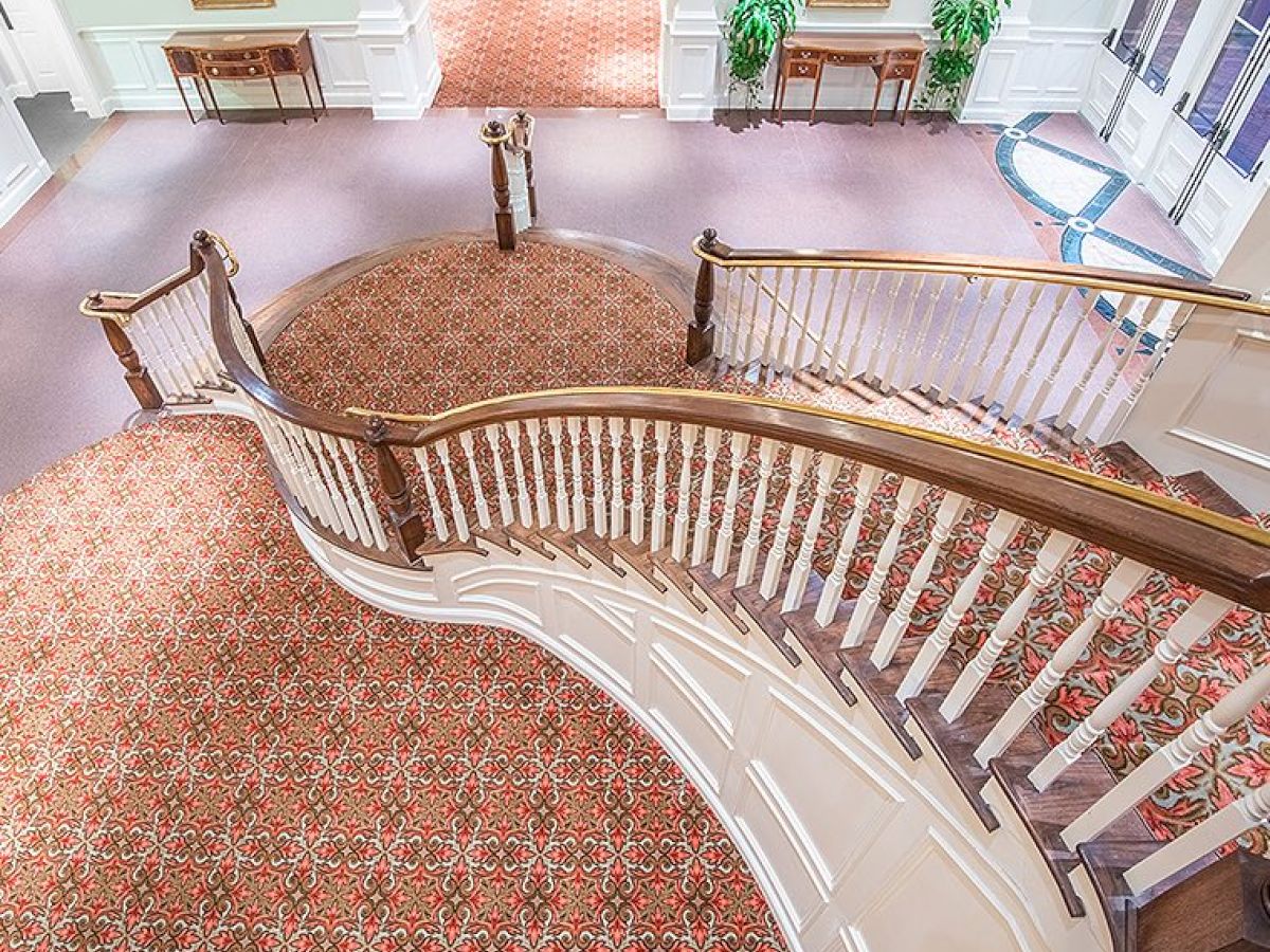 A grand, curved staircase with ornate carpeting and white railings descends to a spacious, carpeted area with tables and decor in an elegant setting.