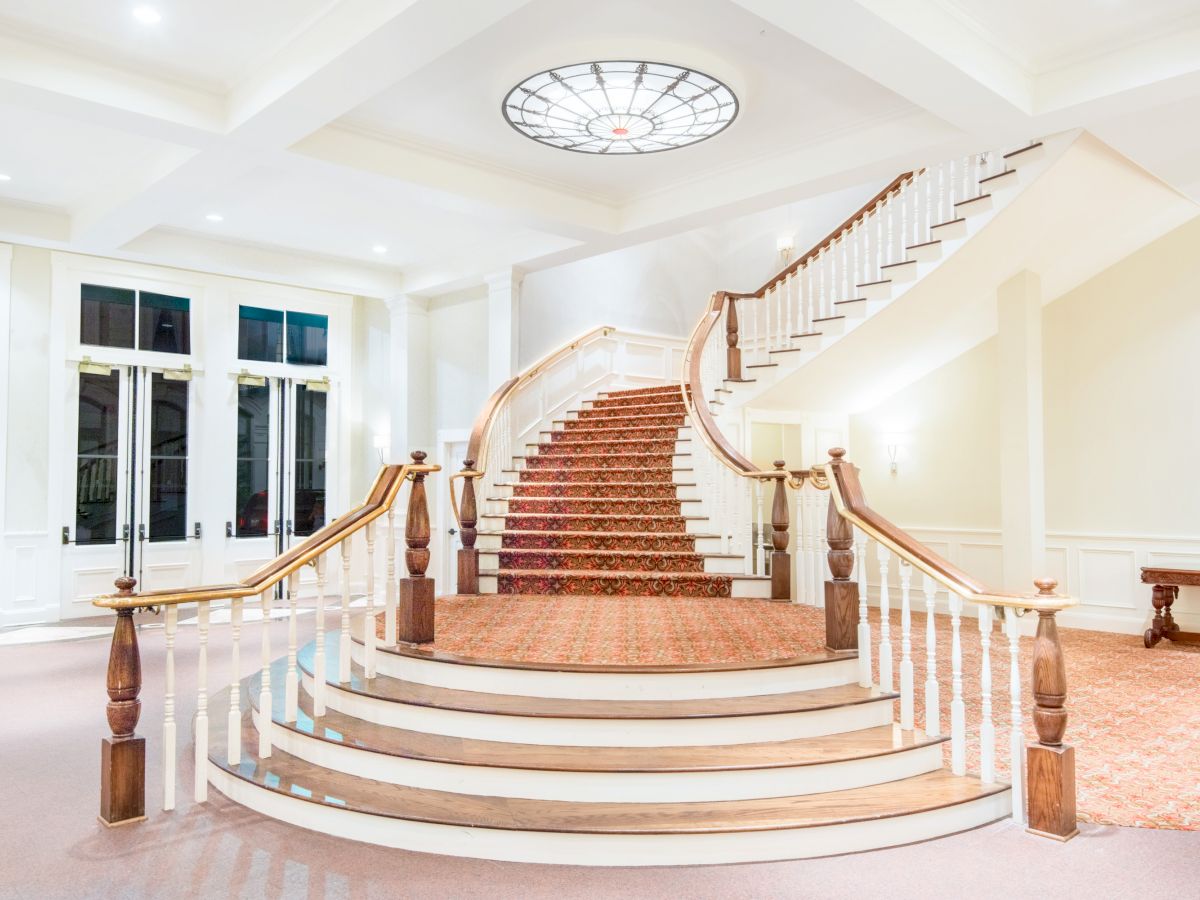 A grand staircase with white railings, wooden handrails, and carpet leads to a mezzanine level in a bright, elegant room with large windows and a ceiling light.