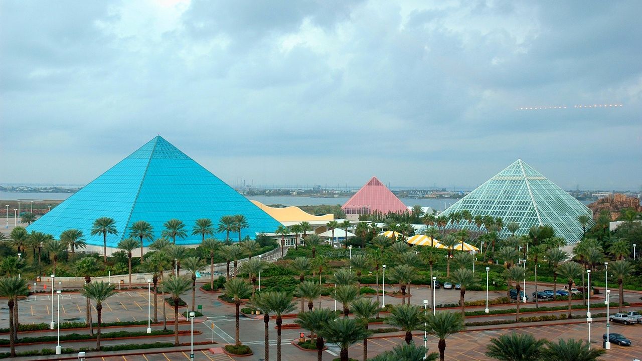 The image shows three pyramidal structures in a landscaped area, with a cloudy sky above and several palm trees in the foreground.