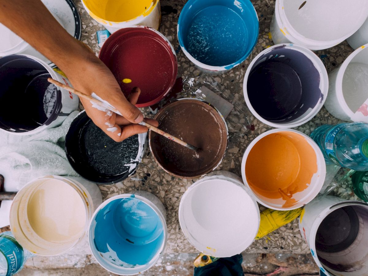 A hand is seen mixing paint in a brown bucket among various colored paint containers, brushes, a hammer, and a couple of water bottles.