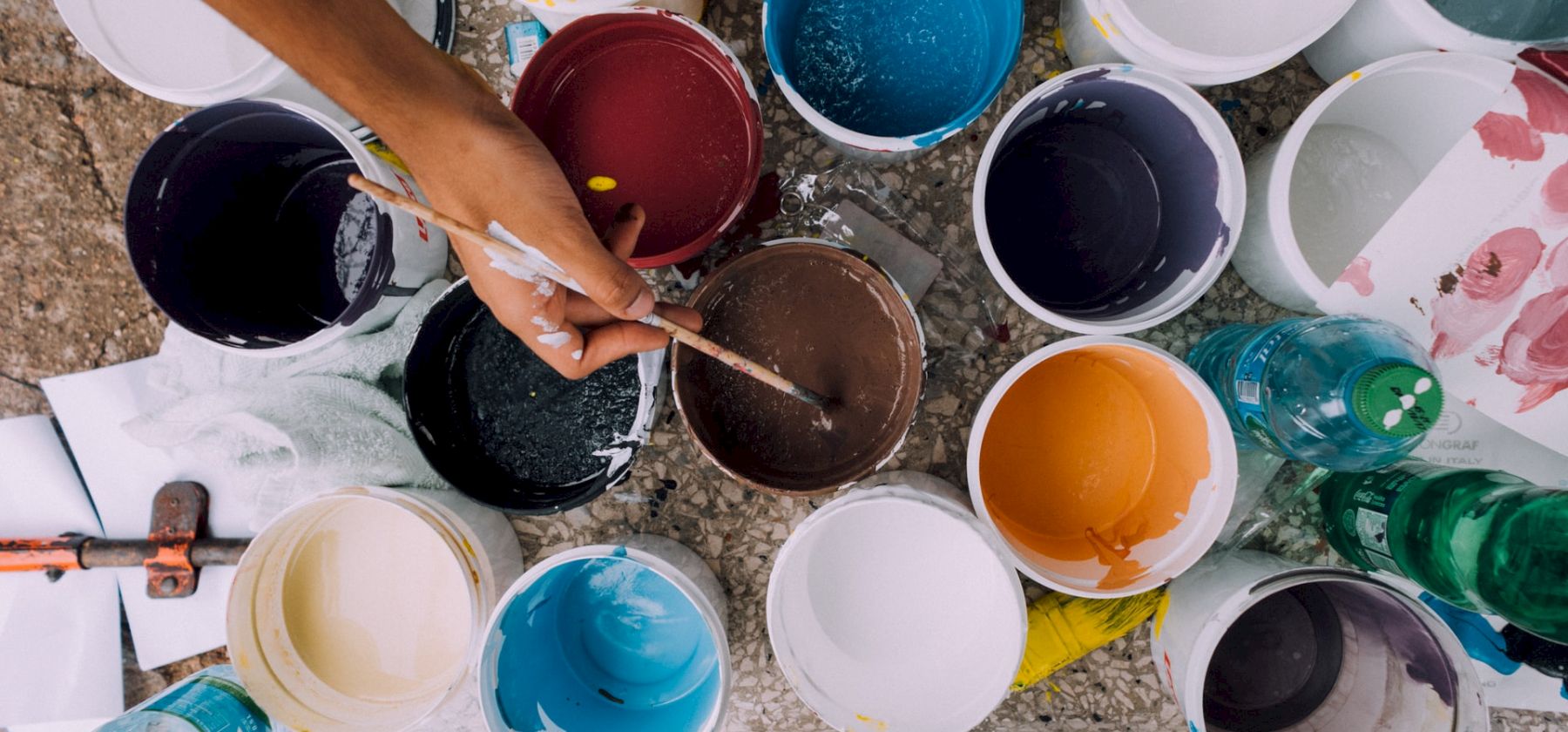 A hand is seen mixing paint in a brown bucket among various colored paint containers, brushes, a hammer, and a couple of water bottles.
