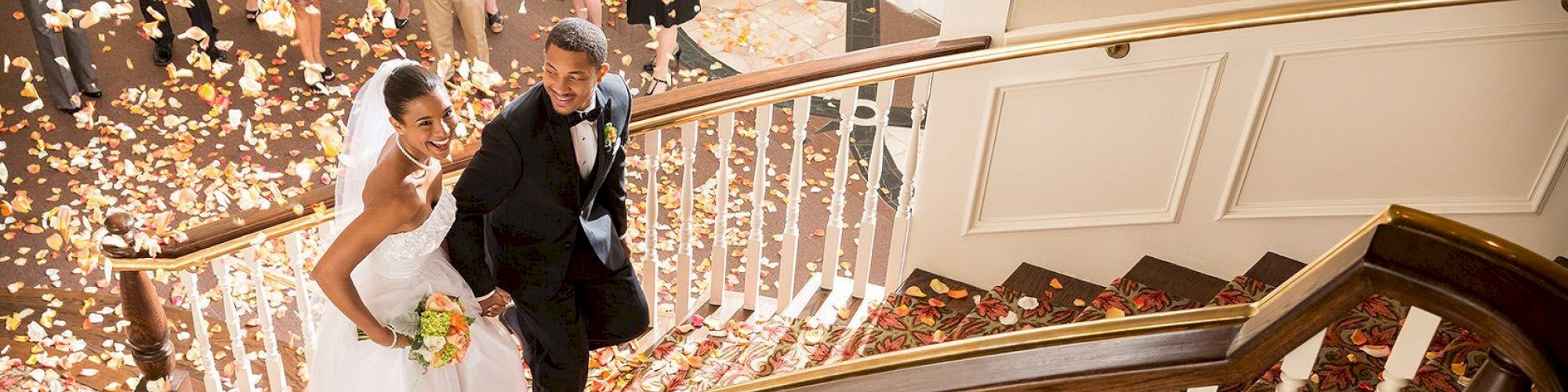 A newlywed couple is walking up a staircase, surrounded by guests and flower petals, while smiling and holding a bouquet.