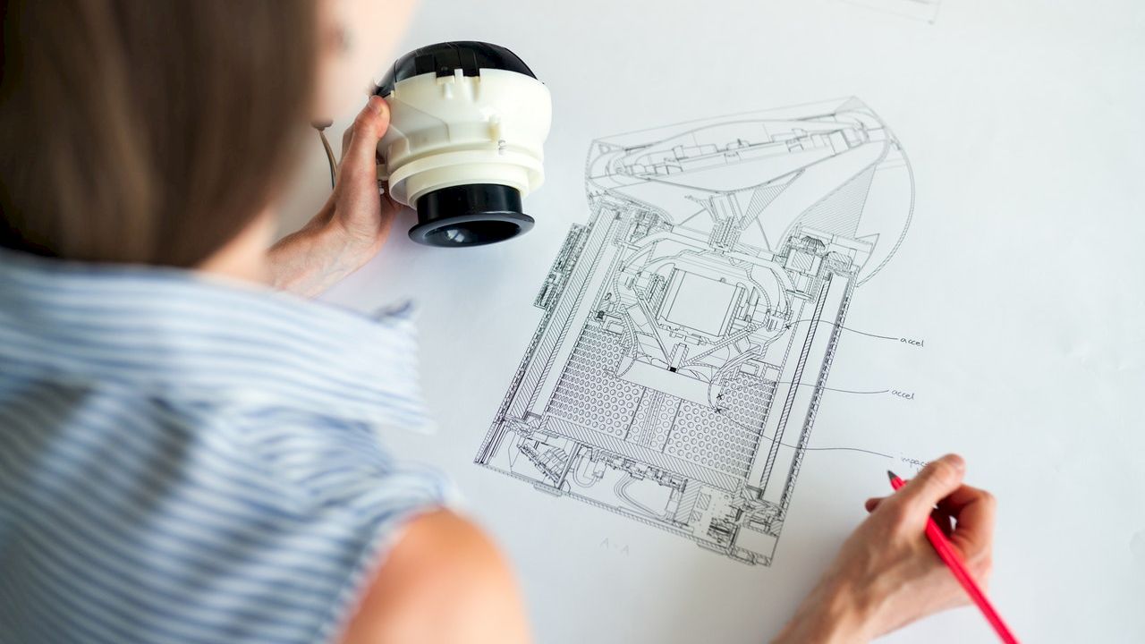 A person is holding a white and black object while looking at technical drawings and using a red pencil to annotate the design on a desk.
