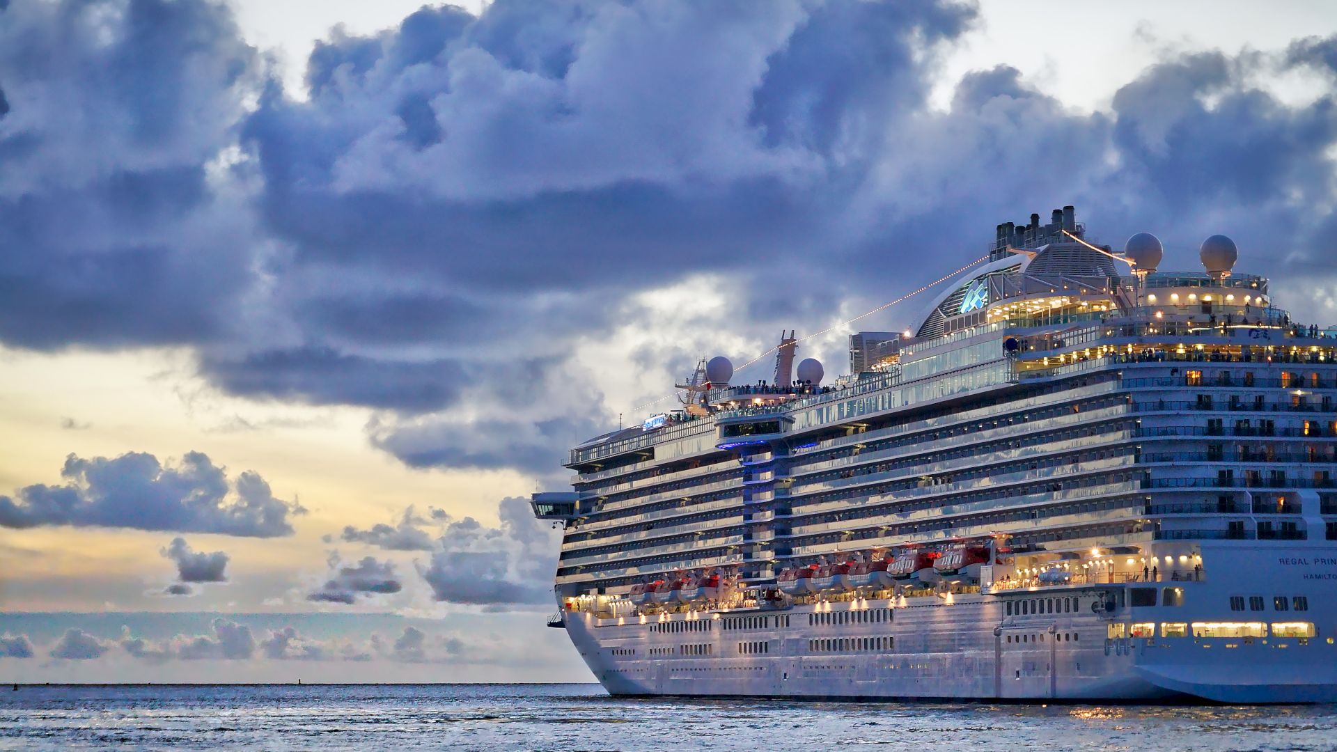 A large cruise ship sails on the ocean under a dramatic, cloudy sky during sunset, with warm sunlight reflecting off the water and the ship.