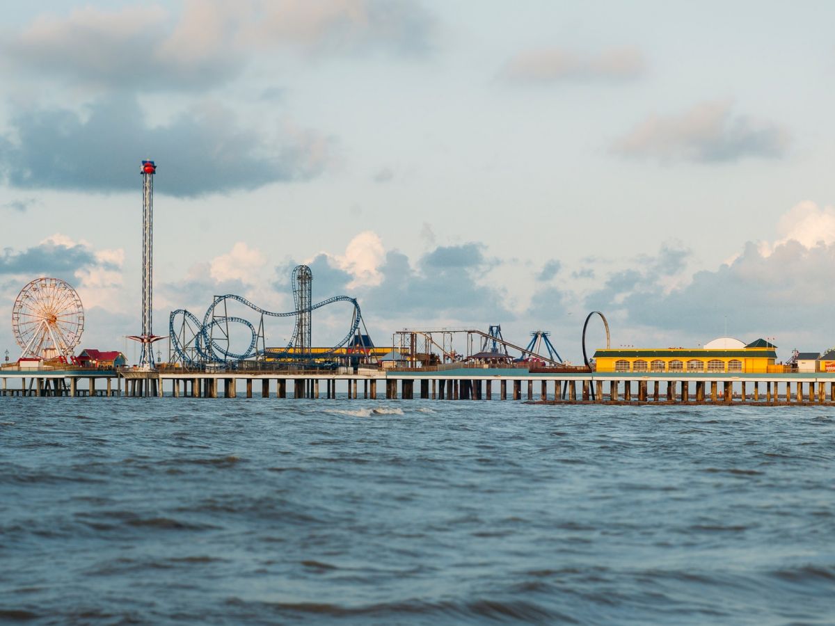 An amusement park over the ocean, featuring a Ferris wheel, roller coasters, and various attractions on a pier, with a cloudy sky backdrop.