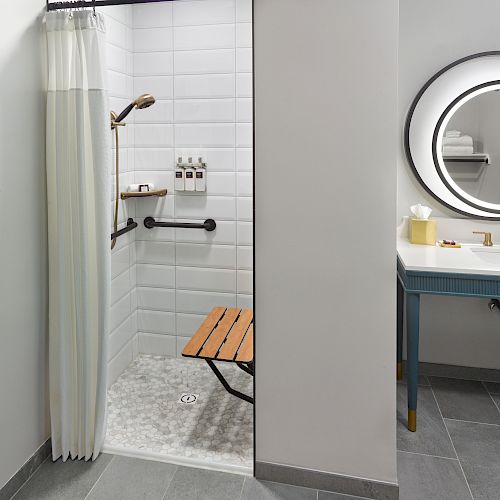 This image shows an accessible bathroom with a shower area featuring a foldable wooden bench and handrails. A sink and circular mirror are adjacent.