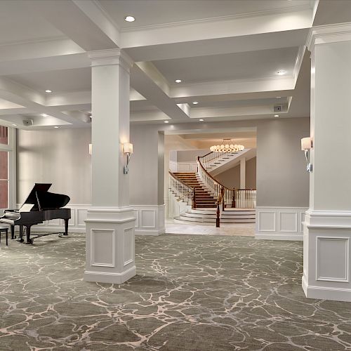 A spacious, elegantly designed room features a grand piano on the left, plush seating on the right, and a grand staircase under a chandelier.