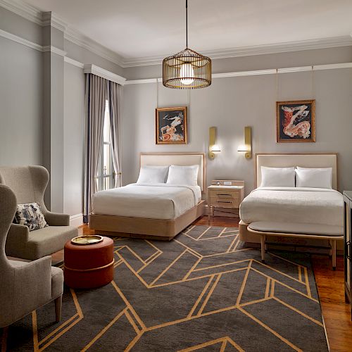 A modern hotel room with two double beds, a chair, a small table, a TV console, wall art, and a geometric-patterned rug.