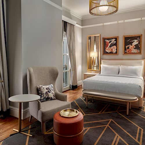 A stylish, modern hotel room with a bed, artistic wall decor, a gray armchair, a round table, an ottoman, and large windows with elegant drapes.