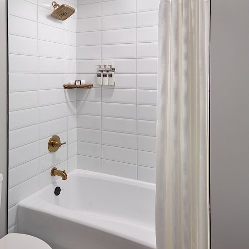 The image shows a clean, white-tiled bathroom with a bathtub, showerhead, wall shelf with toiletries, and a white shower curtain.