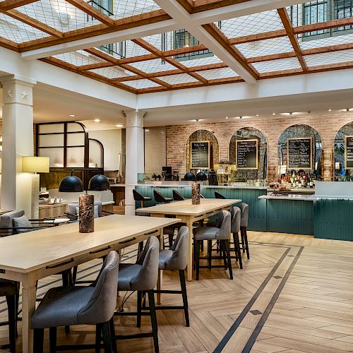 The image shows a modern, well-lit cafe with wooden furniture, a long communal table, high chairs, and a counter area with menu boards.