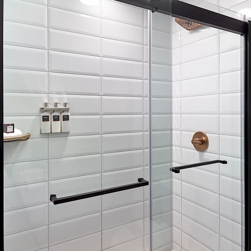 The image shows a modern shower with white subway tiles, glass doors, black handles, a built-in shelf holding toiletries, and a bronze showerhead.