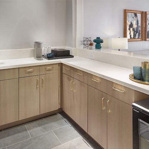 The image shows a sleek, modern kitchen with light wood cabinets, white countertops, a small sink, coffee maker, and a tray with glasses and a carafe.