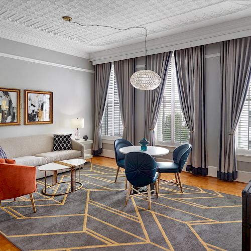 A modern living room features a gray sofa, orange chair, round dining table with blue chairs, patterned rug, wall art, and large windows with gray curtains.