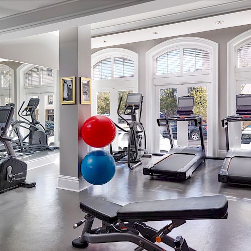 A modern gym with treadmills, exercise bikes, a bench, red and blue exercise balls, and a mirrored wall, well-lit by large windows ending the sentence.