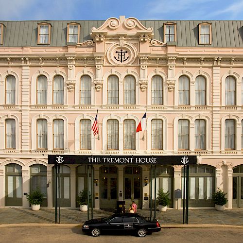 This image shows the grand façade of The Tremont House, a hotel with ornate architectural details, flags, and a black car parked at the entrance.