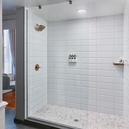 The image shows a modern shower with white tiles, a glass door, wall-mounted shampoo bottles, and gold-colored fixtures, with a room visible outside the shower.