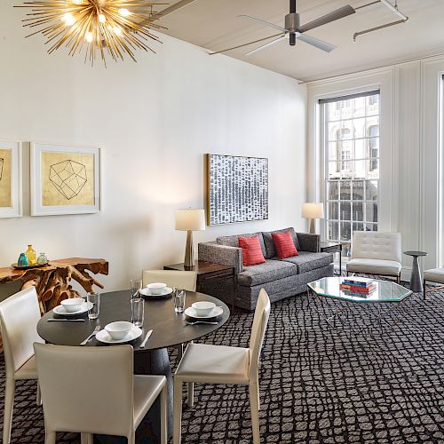 A stylish living room with modern furnishings: dining table, sofa, coffee table, art on the walls, and large windows, lit by a unique chandelier.