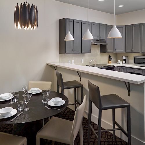 A modern kitchen with gray cabinets, bar seating, and pendant lights next to a dining table set for four with dishes and glassware, ending the sentence.