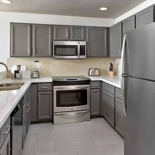 Modern kitchen with gray cabinets, white countertops, stainless steel appliances, and a tiled floor. Includes a microwave, stove, and refrigerator.