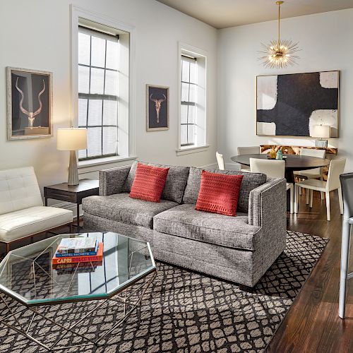 A modern living and dining area with a gray sofa, red pillows, a glass coffee table, stylish decor, bar stools, and large windows.