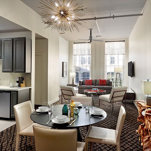 The image shows a modern living space with a kitchen, dining area, and seating area, featuring stylish decor and a unique chandelier.