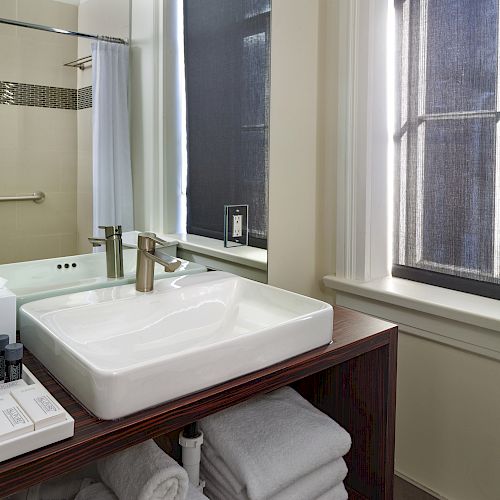 The image shows a modern bathroom with a rectangular sink, a large mirror, toiletries, and a stack of towels on a wooden vanity near a window.