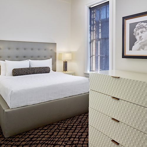 A modern bedroom with a gray upholstered bed, white bedding, nightstands, lamps, a chest of drawers, and framed artwork.