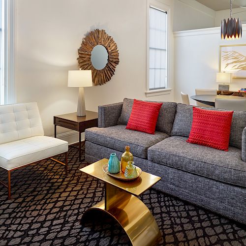 A modern living room with a gray sofa, red pillows, white chair, gold coffee table, lamp, decorative mirror, abstract art, and dining table.