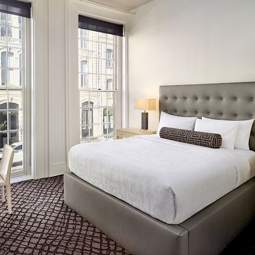 A modern hotel room features a neatly made bed with a headboard, two side tables with lamps, a window with tall curtains, and a desk and chair.