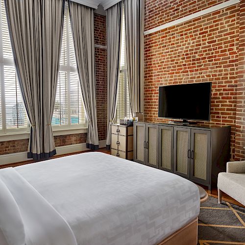 A well-lit bedroom with brick walls, large windows, curtains, a bed, TV on a cabinet, and a cushioned chair.