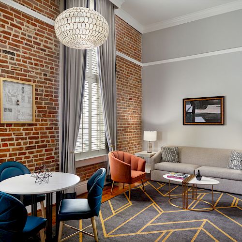 This is a modern living room with brick walls, gray couch, armchairs, a round dining table with blue chairs, a geometric rug, and large windows.