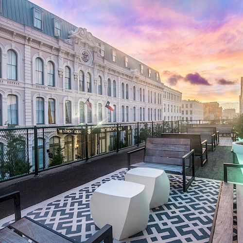 A rooftop terrace with modern seating and potted plants overlooks classic buildings at sunset.