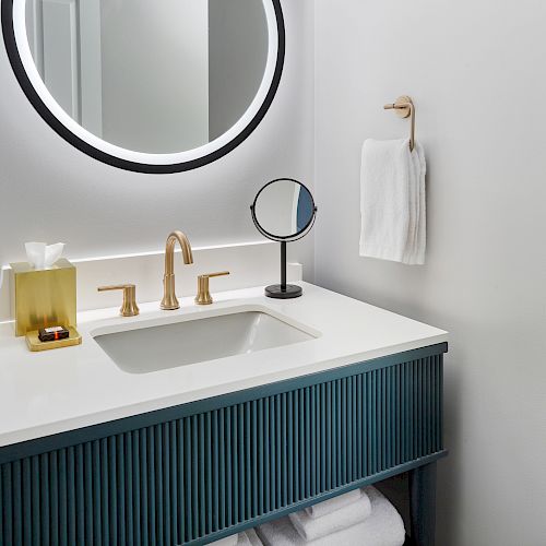 This image shows a modern bathroom sink with gold fixtures, a round illuminated mirror, a tissue box, a small round mirror, and a towel hanging on the wall.