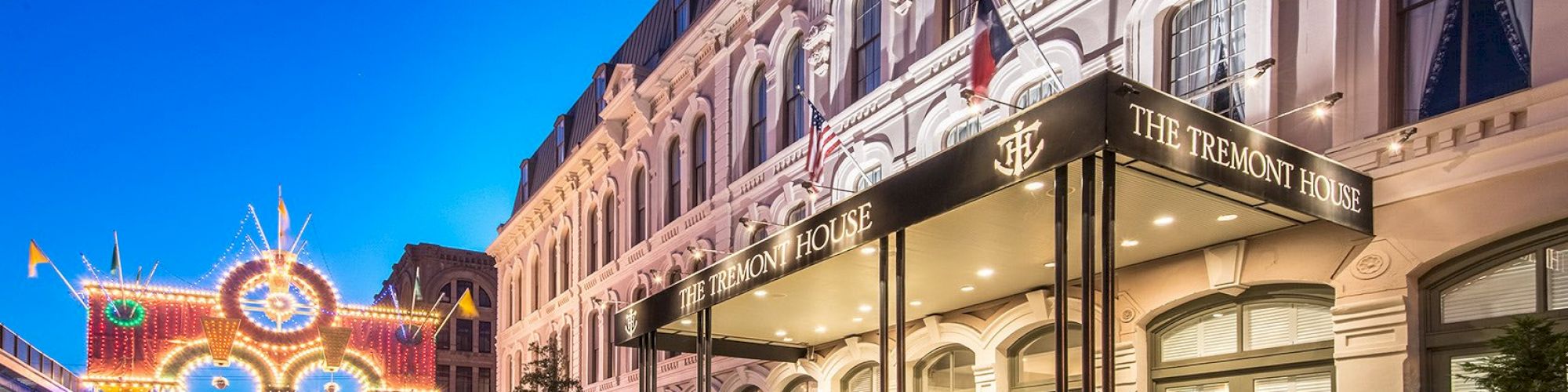 A grand historic building with "The Tremont House" signage is adorned with lights at dusk, featuring a festive archway down the street.