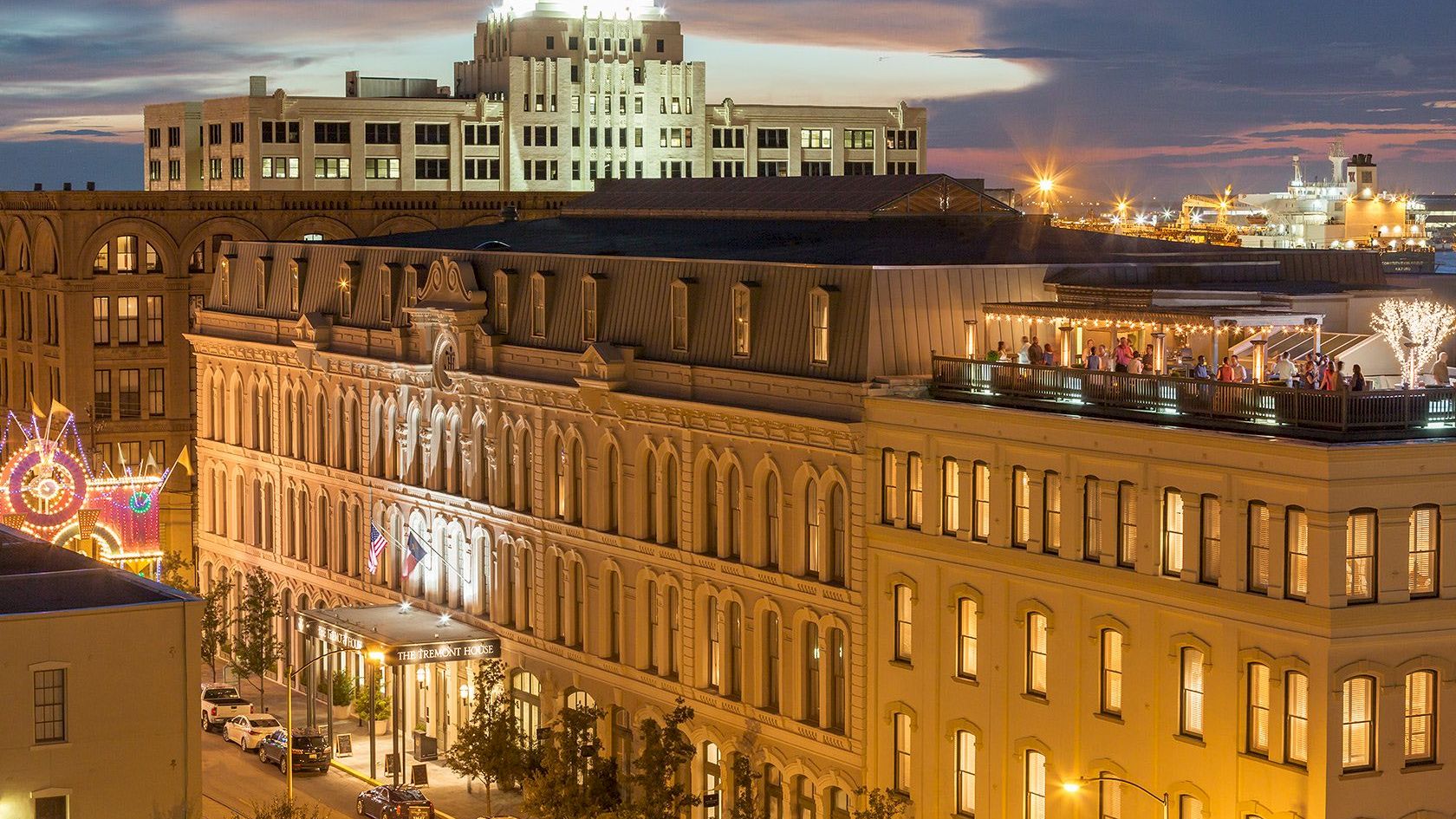 The image shows a cityscape at dusk with a historic multi-story building in the foreground, adorned with lights.