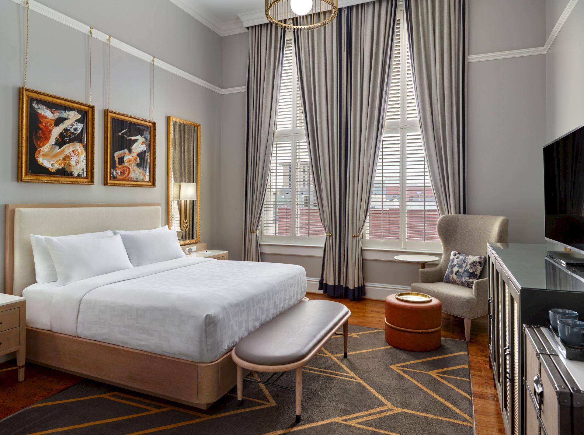 A modern hotel room with a large bed, bedside tables, hanging art, and large windows with curtains. There's also a TV, chair, and small table.