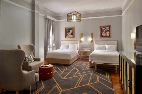 A hotel room with two beds, wall art, a geometric carpet, an armchair, a small table, and a TV on a cabinet.